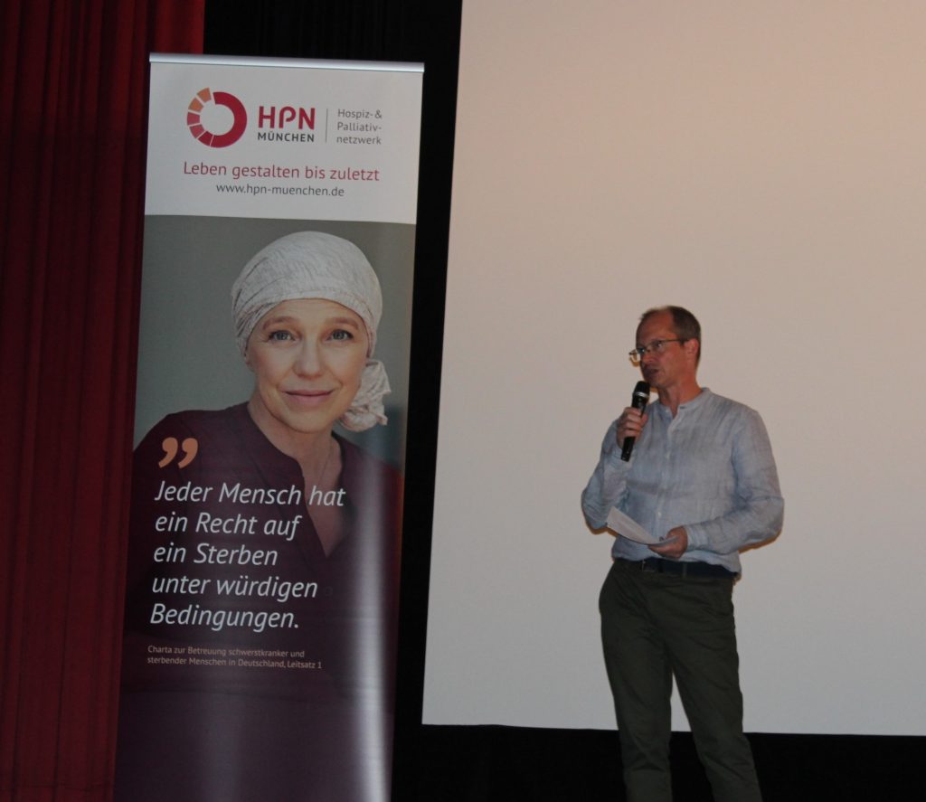 Impressionen der 1. Münchner Hospiz- und Palliativwoche im Oktober 2024. Ein Herr hält einen Vortrag, links neben ihm ein Plakat mit dem Text der Charta zur Betreuung schwerstkranker und sterbender Menschen.