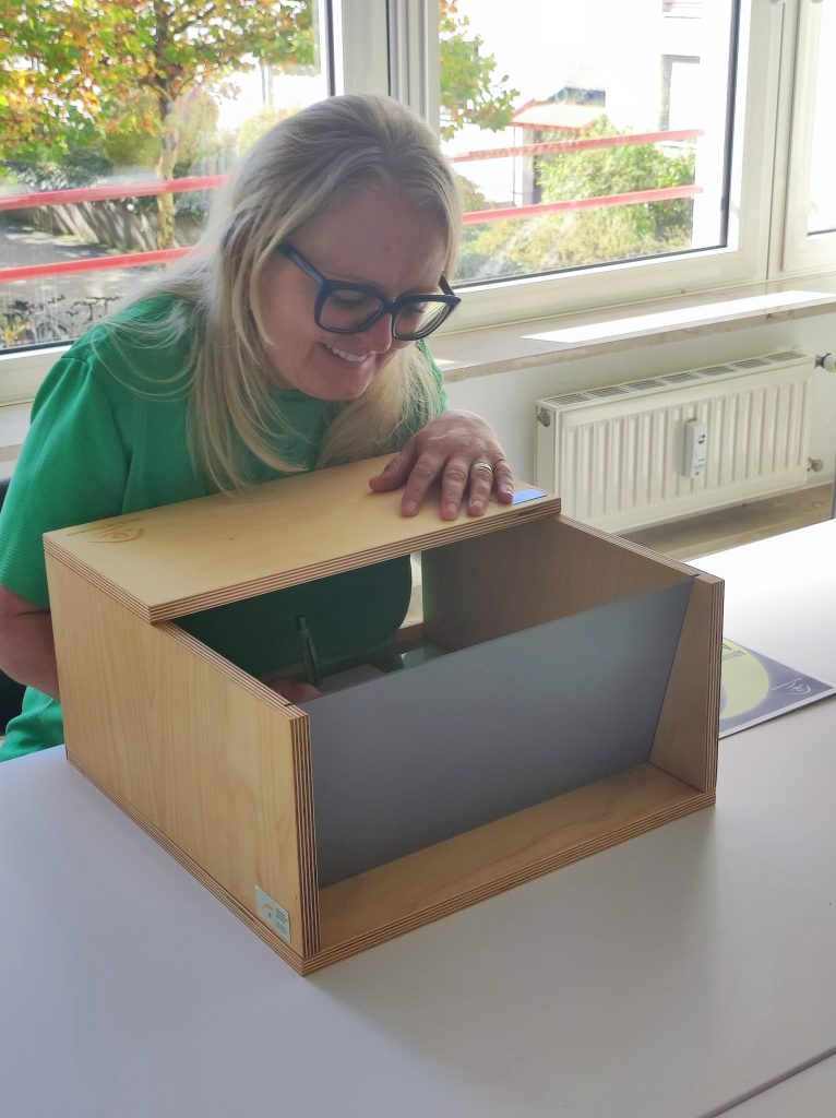 Impressionen der 1. Münchner Hospiz- und Palliativwoche im Oktober 2024. Besucher sitzen in einem Raum und sehen sich eine Präsentation oder Vortrag an. Ein Frau füllt ein Formular in einem blick-schützendem Kasten aus.