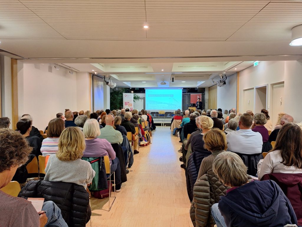 Impressionen der 1. Münchner Hospiz- und Palliativwoche im Oktober 2024. Besucher sitzen in einem Raum und sehen sich eine Präsentation oder Vortrag an.